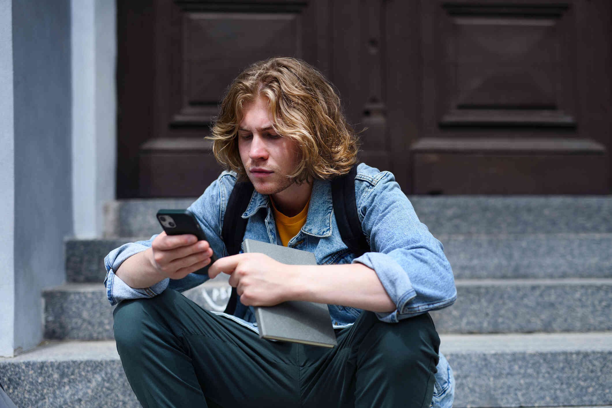 A man in a jean jacket and bakc pack sits on a cement step outside and looks at the cellphone in his hand.
